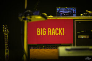 Red rack panel with bold yellow 'BIG RACK!' text on stage, guitar to the left and audio equipment below it.