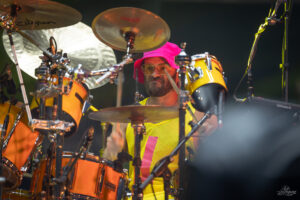 Drummer wearing a bright pink hat and yellow sleeveless shirt plays an orange drum set on stage.