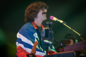 Musician with a mustache and glasses singing into a pink microphone on stage, wearing a retro blue, white, and red striped jacket.