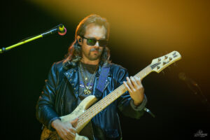 Male bass guitarist wearing sunglasses and a black leather jacket playing a five‑string bass on stage under a warm yellow spotlight.