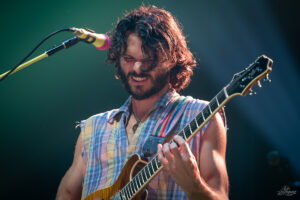 Male guitarist with shoulder-length hair and a beard plays an electric guitar on stage, smiling while performing into a microphone.