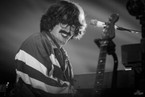 Male musician with shaggy hair and mustache, wearing a striped jacket, playing a keyboard on stage with a microphone nearby (black and white).