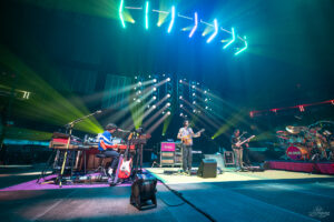Live band on a lit stage with keyboardist, guitarist, bassist, and drummer performing under colorful spotlights.