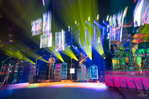 Band performs on a brightly lit concert stage with yellow and blue spotlights and oversized video screens above them.