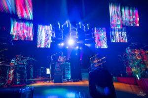 Live concert scene with blue stage lighting, musicians playing guitars, and colorful LED screens overhead illuminating the band.