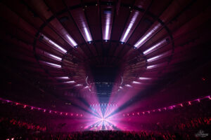 Large indoor concert arena with a band on stage and pink-purple beam lights illuminating the crowd.