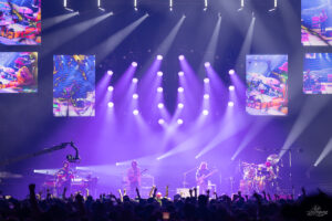 Live concert on a large stage with purple spotlight beams and screens above, four musicians playing guitars/drums while the crowd raises hands.