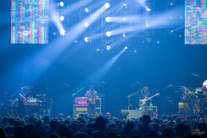 Live concert stage with four musicians performing under blue lights, a crowded audience in the foreground.