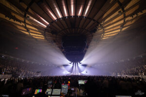 Interior of Madison Square Garden during a concert: a full crowd surrounds a central stage with powerful white spotlights radiating outward.