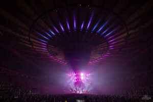 Indoor concert arena filled with a large crowd, vibrant purple stage lights and a central screen above performers.] ,