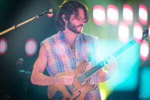 Guitarist with a beard wearing a plaid sleeveless shirt, playing a wooden electric guitar on stage with colorful lights behind him.