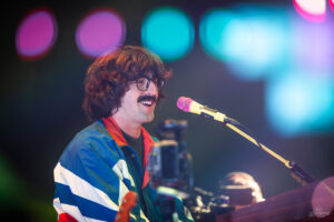 Singer with a mustache and round glasses performs on a stage, wearing a blue, white, and red jacket in front of colorful lights.