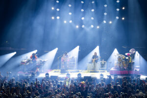 Band performing on a well-lit stage with guitarists and a drummer, bright spotlights overhead, crowded audience hands in the air in front of them toward a live show.