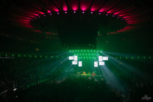Wide shot of a packed arena at night: a concert stage with green lights and hanging screens above the audience.
