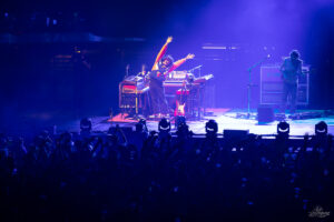 Musician on stage with arms raised, surrounded by keyboards and guitars, blue spotlight over a cheering crowd on the arena floor.
