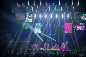 Concert stage with a four-piece band performing under bright multi-colored lights, surrounded by large video screens showing abstract visuals and crowd in the foreground.