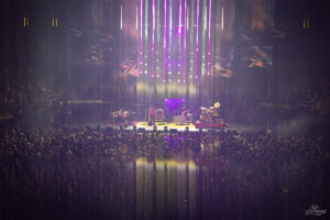 Concert crowd faces a lit stage with a band performing under purple vertical lights and haze.