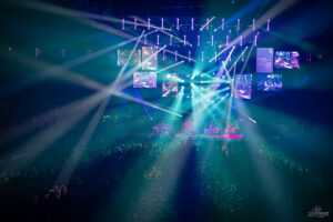 Large indoor concert venue with a full band performing on stage under colorful laser lights and multiple screens above the crowd.