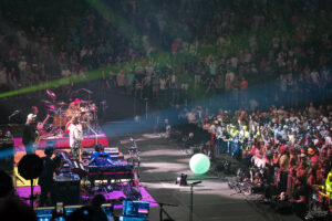 Band performing on a lit stage to a packed crowd with colorful spotlights and a glowing ball on the floor.
