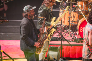 Bearded saxophonist wearing a patterned cap plays on stage next to a guitarist, with a drum kit visible in the background on a red-carpeted stage.