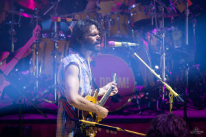 Male guitarist with a beard sings into a pink microphone on a colorful stage with drums in the background.
