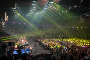 Large indoor concert with a band performing on stage and bright green and yellow lights illuminating a packed arena audience.