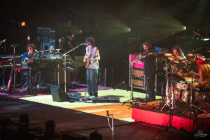 A rock band performs on stage: keyboardist left, guitarist center, singer at microphone, and drummer on the right under colorful spotlights.
