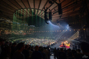 Large indoor concert arena filled with standing crowds facing a brightly lit stage with musicians performing.