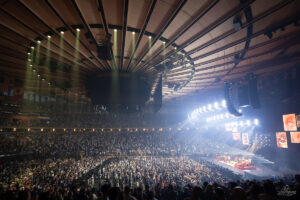 Crowd fills a large indoor arena as a concert stage lights up, with a circular overhead screen and hanging rigging overhead.