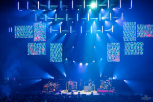 Live concert on a blue-lit stage with a band playing and psychedelic video panels above all around the stage.