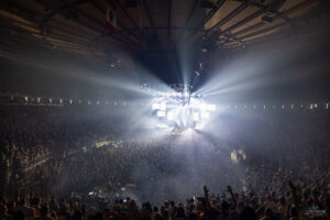 Massive indoor concert with a brightly lit stage and hanging screens, crowd cheering in a packed arena.