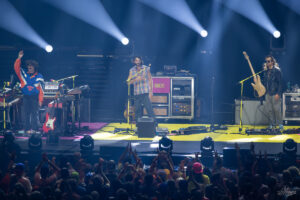 Three musicians perform on a stage with keyboards, guitar, and bass under bright spotlights as the cheering crowd watches.