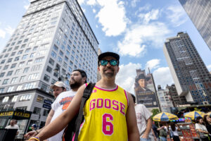 Man wearing a yellow Goose jersey with the number 6 walks toward the camera in a busy city street, tall buildings around him and blue sky above.