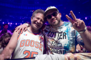Two men posing together at a lively indoor event, one in a GOIGE GOOSE 25 basketball jersey and the other in a tie-dye shirt, both smiling with arms around each other and making a peace sign.