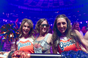Three smiling women at a concert, two wearing orange Goose shirts and holding colorful pom-poms, under blue stage lighting.