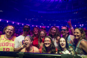 A group of smiling fans at a concert barrier under purple stage lights, posing for a photo together.