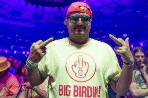 Man at a concert wearing sunglasses and a pink cap, flashing middle fingers with a cream T-shirt reading 'BIG BIRD IN!' amid colorful club lighting.