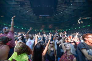 A large, excited crowd at a concert in an arena, many fans raising their hands and cheering under blue arena lighting.
