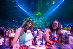 Concert crowd at a goose-themed event, two smiling women in white and orange Goose shirts near the barrier under blue and green stage lights.