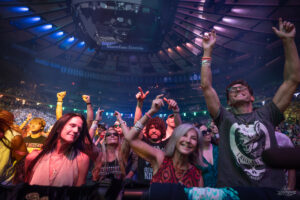 Energetic concert crowd near the stage, fans raise their hands and cheer under colorful arena lights.