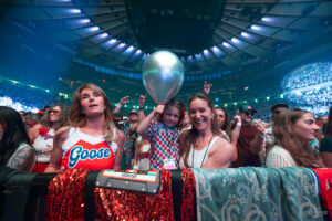 Front-row concert fans: a child with a silver balloon flanked by two smiling women, at a Goose show with blue arena lighting.
