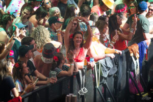 Woman in a red bikini top forms a heart shape with her arms at a sunlit festival barrier, surrounded by cheering crowd.