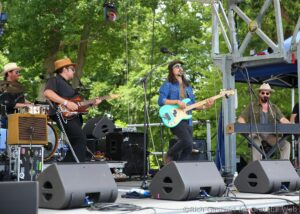 Live band performing outdoors on a sunny stage with guitarists, a bassist, and a drummer; green trees in the background.