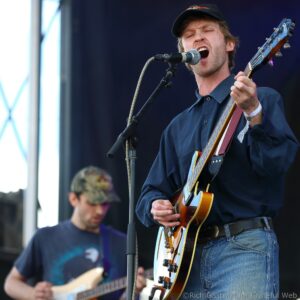 Male guitarist singing into a microphone while playing an electric guitar on stage.