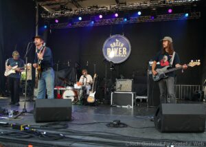 A four-person rock band performing on an outdoor stage with guitars, drums, and a large circular 'Green River Festival' sign in the background.