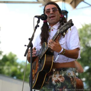 Male guitarist singing into a microphone outdoors, long hair, pink patterned sunglasses, wearing a light shirt and playing an acoustic guitar.