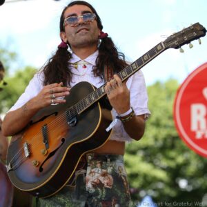 Guitarist with long hair and patterned pants, wearing sunglasses and a white shirt, playing an acoustic-electric guitar outdoors at a festival.