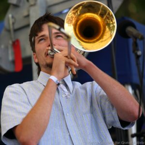 Close-up of a man playing a brass trumpet, lips on the mouthpiece, wearing a light striped shirt.