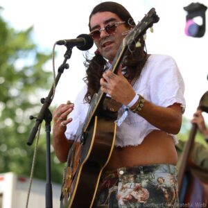 Male guitarist with long hair and patterned sunglasses sings into a microphone while playing an acoustic guitar outdoors on stage.