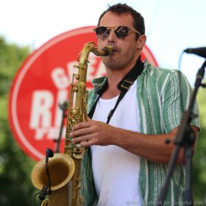 Musician playing a saxophone outdoors, wearing sunglasses and a green striped shirt, with a red circular backdrop behind him.